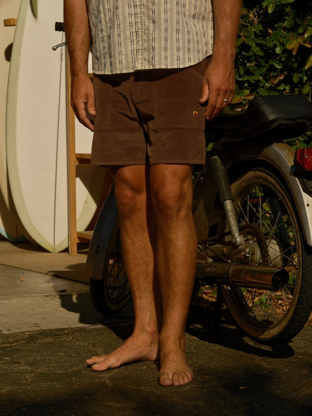 A barefoot man in Mollusk - Men's Shroom Short / Chocolate stands next to a bike and surfboard, with greenery and sunlight casting shadows in the background.
