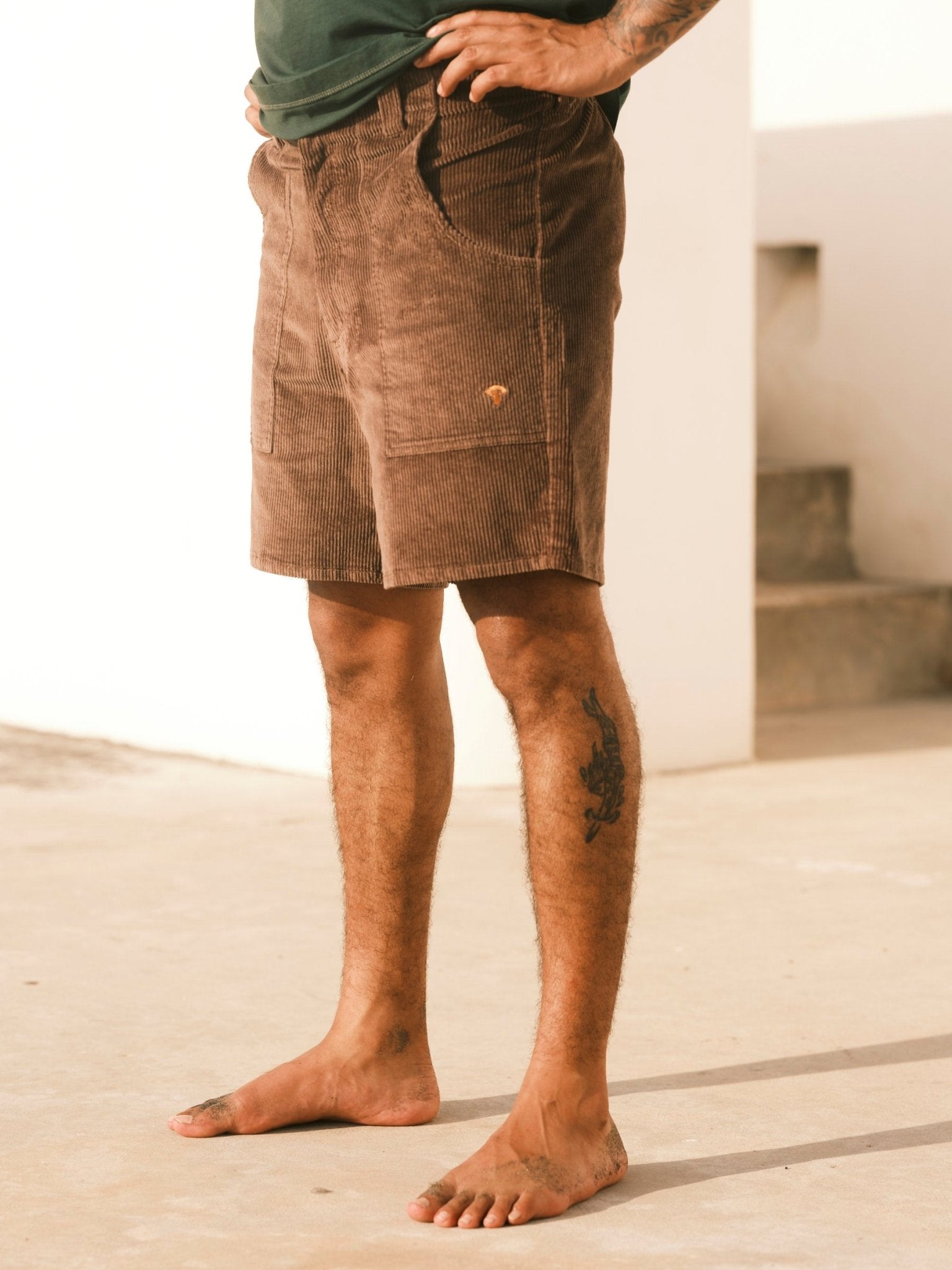A person stands barefoot outdoors in Mollusk Men's Shroom Short (Chocolate) with a dark green shirt, showing a tattoo on their left calf, one hand in a pocket, against a neutral background and staircase.