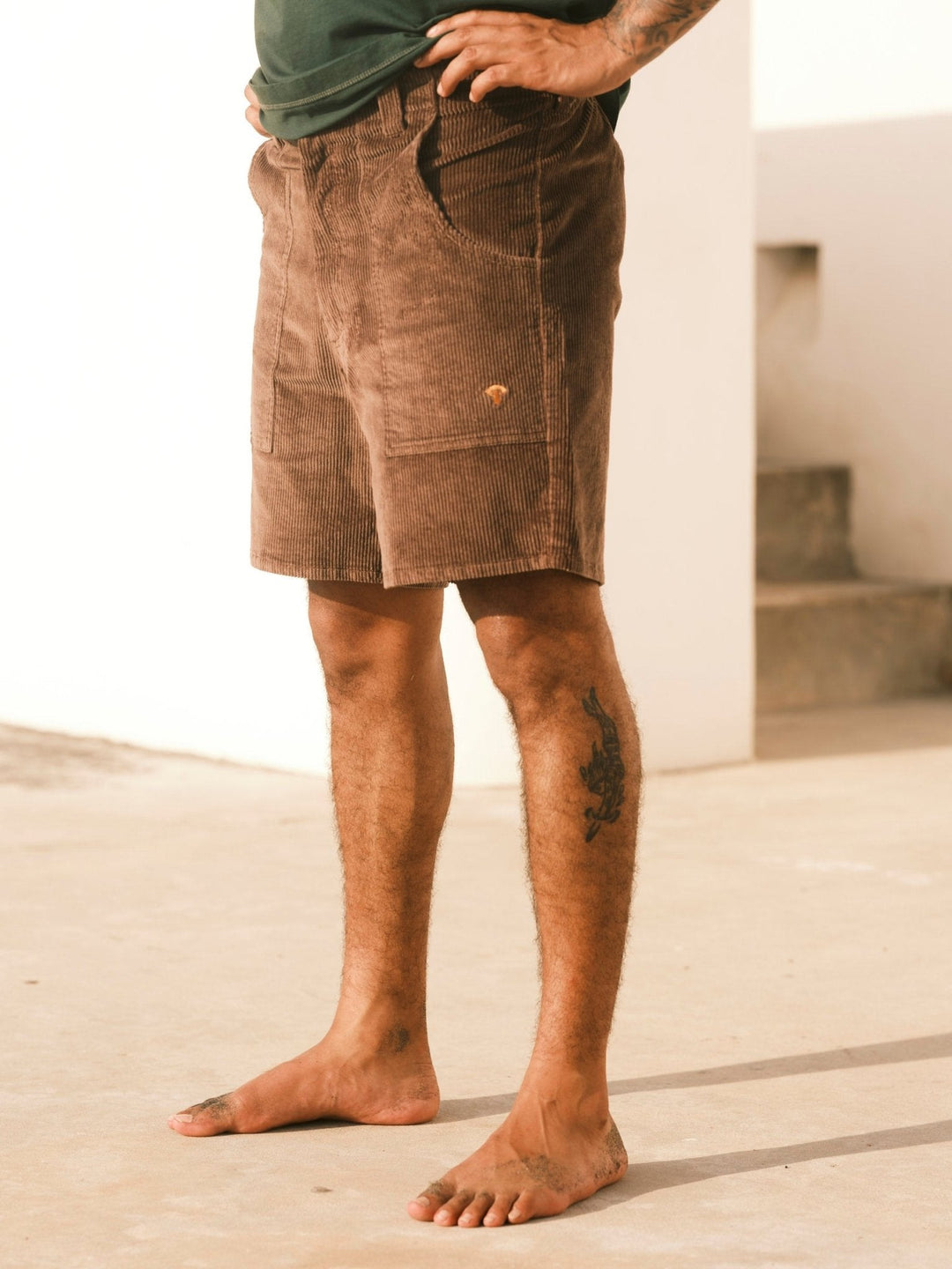 A person stands barefoot outdoors in Mollusk Men's Shroom Short (Chocolate) with a dark green shirt, showing a tattoo on their left calf, one hand in a pocket, against a neutral background and staircase.