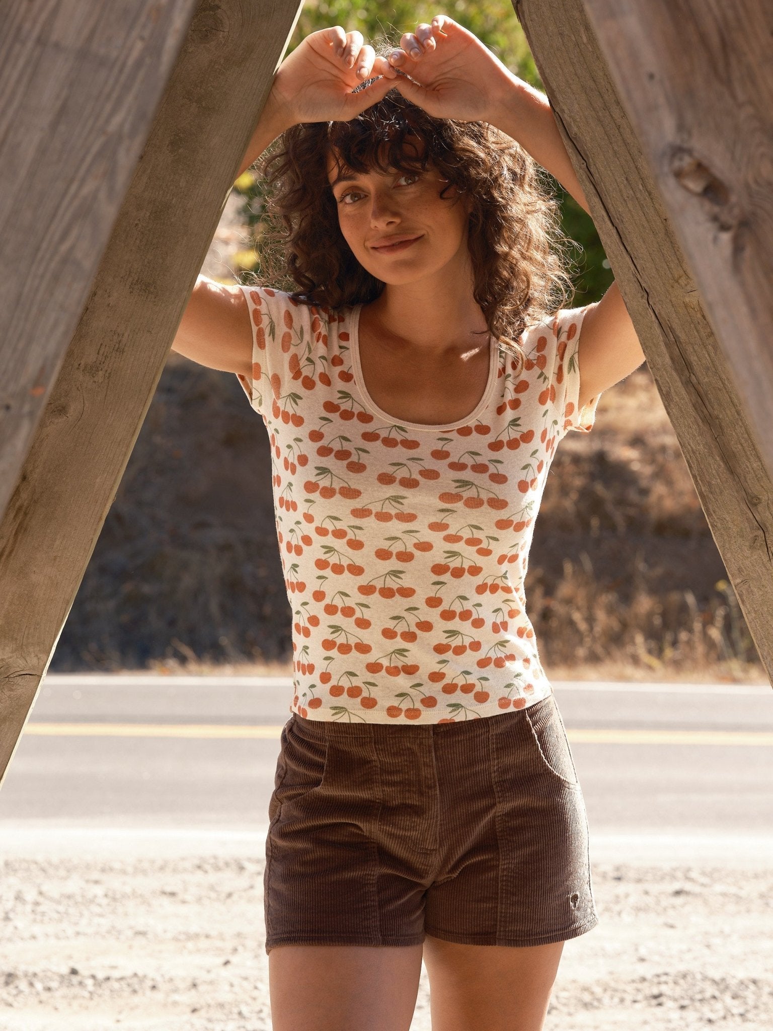 A woman with curly hair stands outdoors between wooden beams, smiling. She wears a white T-shirt with cherries and Mollusk - Women's Shroom Short in Chocolate by Mollusk, with a road and dry grass in the background.