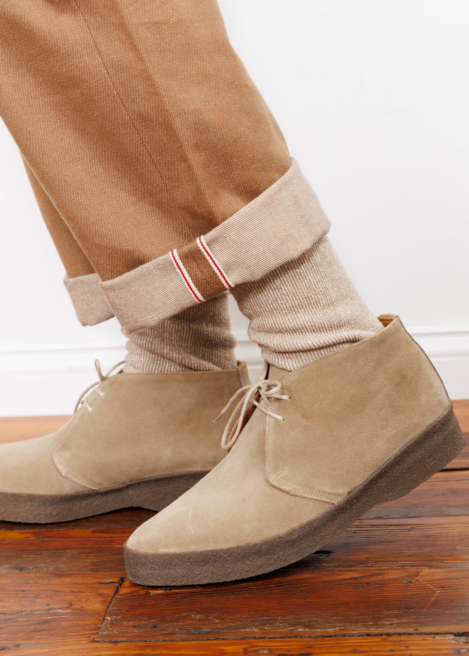 A person wearing American Trench Sunday Sock in khaki, light brown suede desert boots, and tan cuffed pants with red-striped cuffs stands on a polished wooden floor against a white wall.