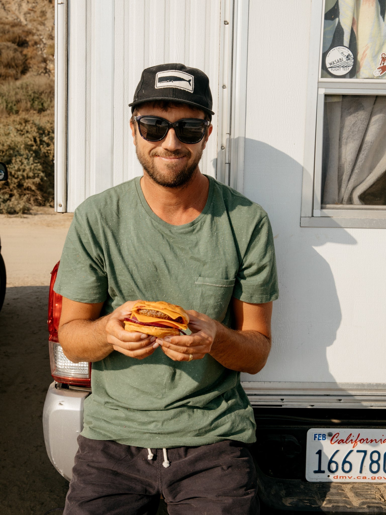 A man in a Mollusk Whale Patch Hat in black smiles and holds a sandwich while sitting on the back of a vehicle with a California license plate.