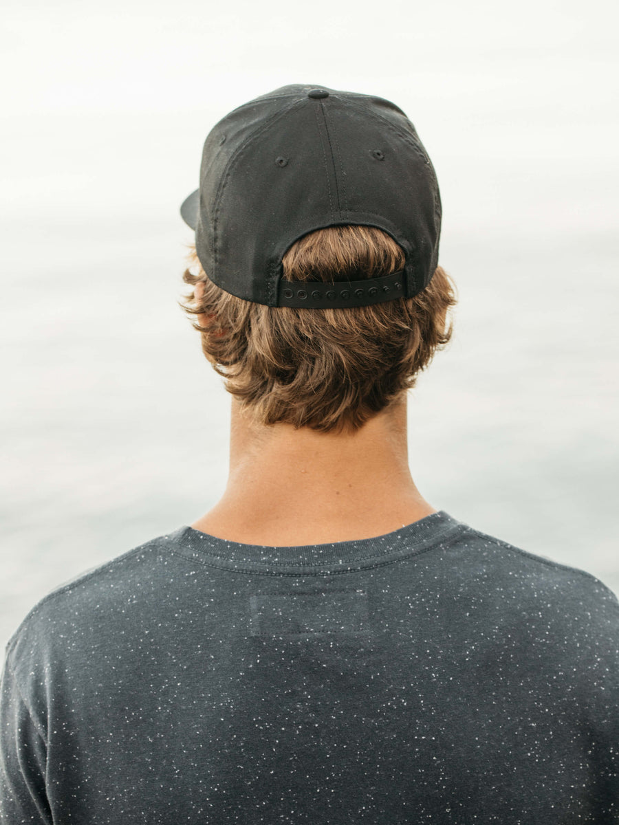 A person with light brown hair is seen from behind, wearing the Mollusk Whale Patch Hat in black and a dark speckled 100% cotton T-shirt, standing before a softly blurred light background.