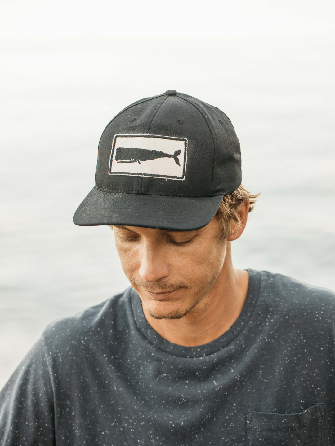 A man in a navy speckled 100% cotton shirt wears the Mollusk - Whale Patch Hat / Black by Mollusk, featuring a white whale patch. He looks down with a neutral expression. The oceanic background is blurred.