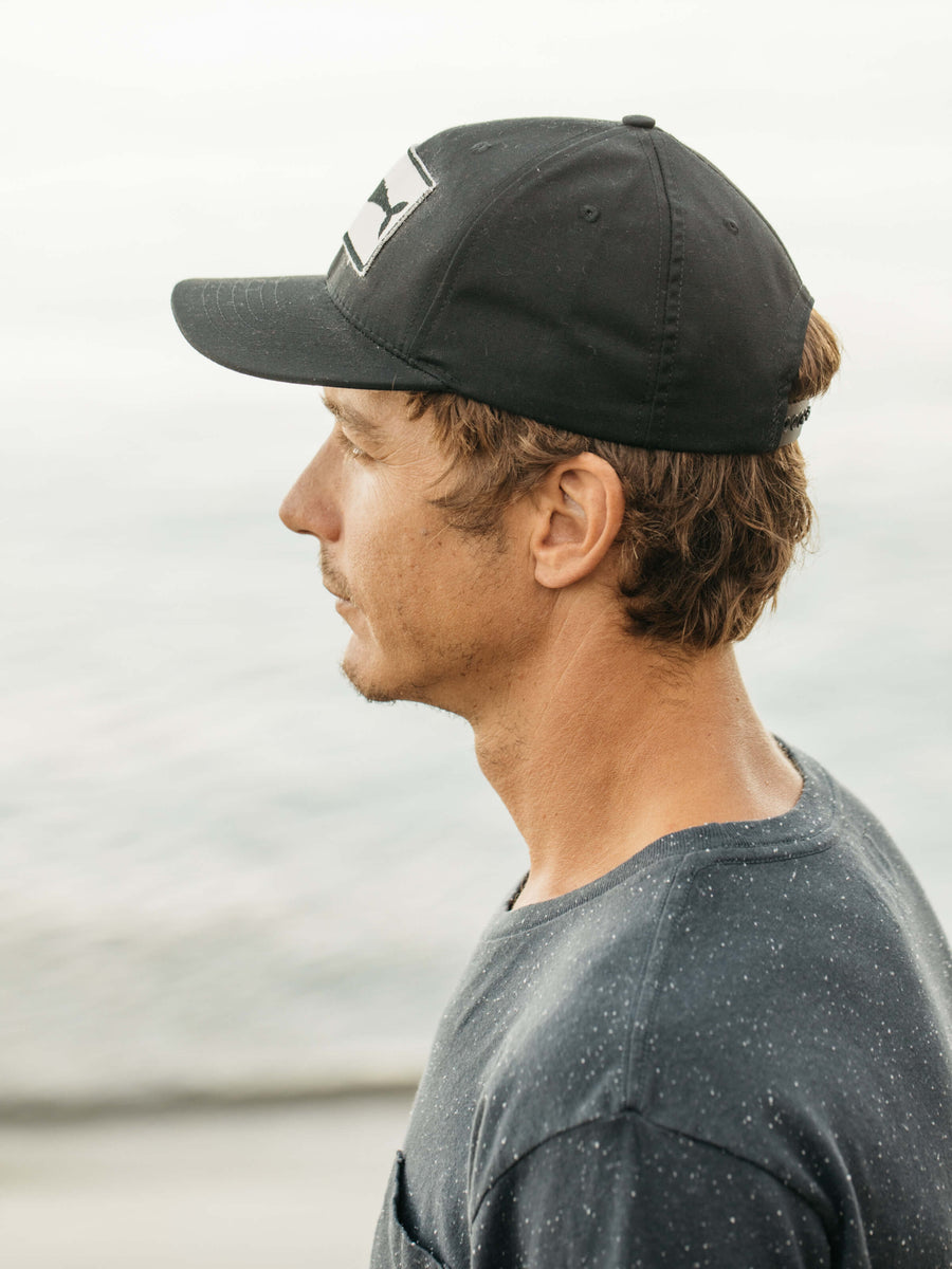 A man with light brown hair is wearing the Mollusk Whale Patch Hat in black and a dark speckled cotton shirt, gazing left in profile against a blurred, oceanic background.