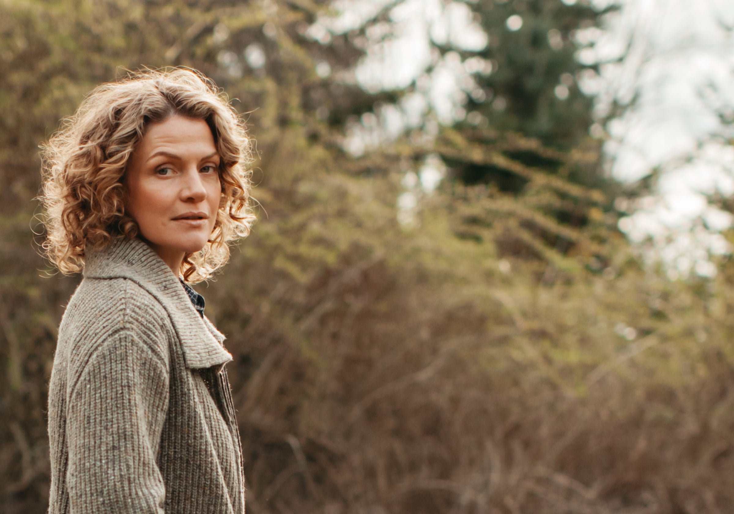 A person with curly hair wearing a textured coat stands outdoors, looking over their shoulder. The background is blurred with trees and shrubs, suggesting a natural setting.
