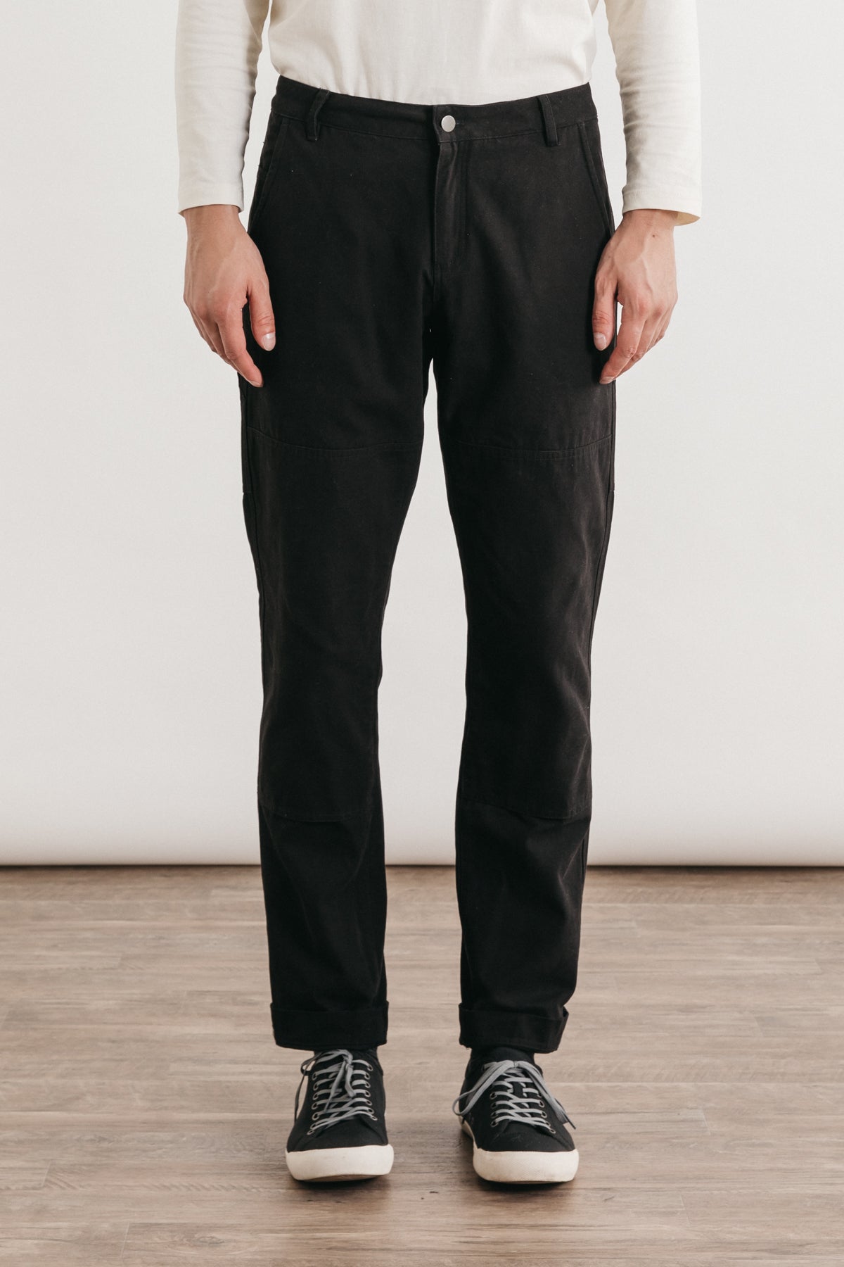 A person indoors, shown from the shoulders down, wears Bridge & Burn’s Wallace Utility Pant in black canvas, paired with a white long-sleeve shirt and black sneakers with white soles.