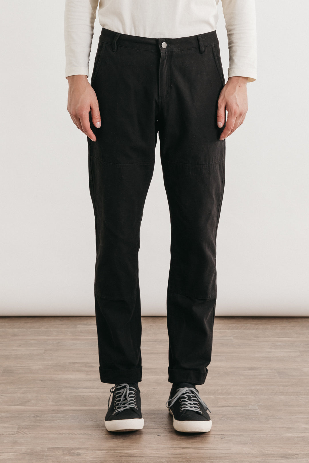 A person indoors, shown from the shoulders down, wears Bridge & Burn’s Wallace Utility Pant in black canvas, paired with a white long-sleeve shirt and black sneakers with white soles.