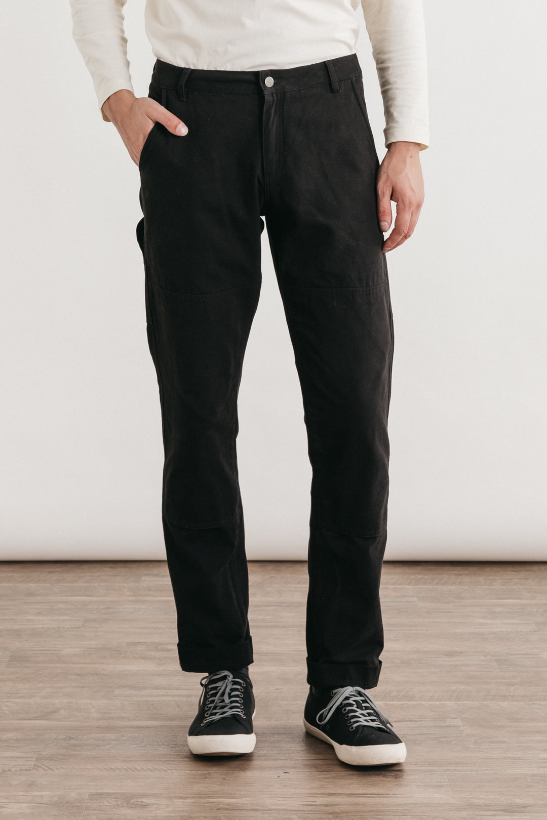 A person stands on a wooden floor with one hand in their pocket, wearing a white long-sleeve shirt and Bridge & Burn’s Wallace Utility Pant in Black Canvas; the image is cropped at the shoulders, keeping their face unseen.