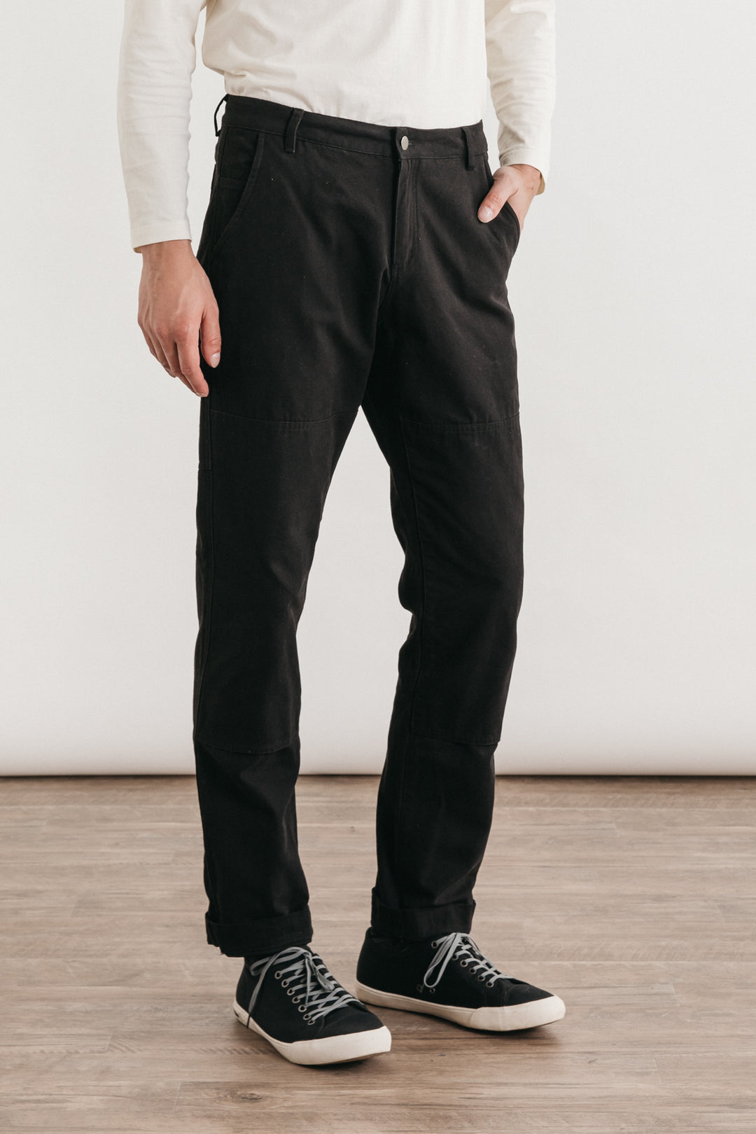 Indoors on a wood floor, a person wears Bridge & Burn’s Wallace Utility Pant in black canvas, paired with a white long-sleeve shirt and black sneakers. Their left hand is in their pocket; their head is out of frame.