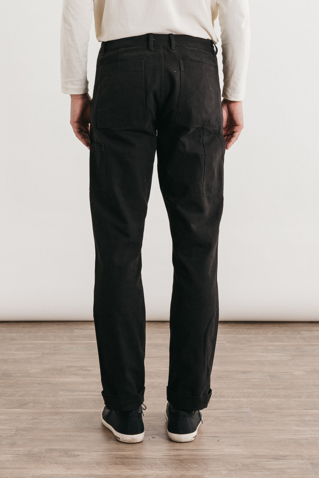A person stands on a wooden floor, facing away, wearing Bridge & Burn’s Wallace Utility Pant in black canvas, a white long-sleeve shirt, and black shoes against a plain light background.