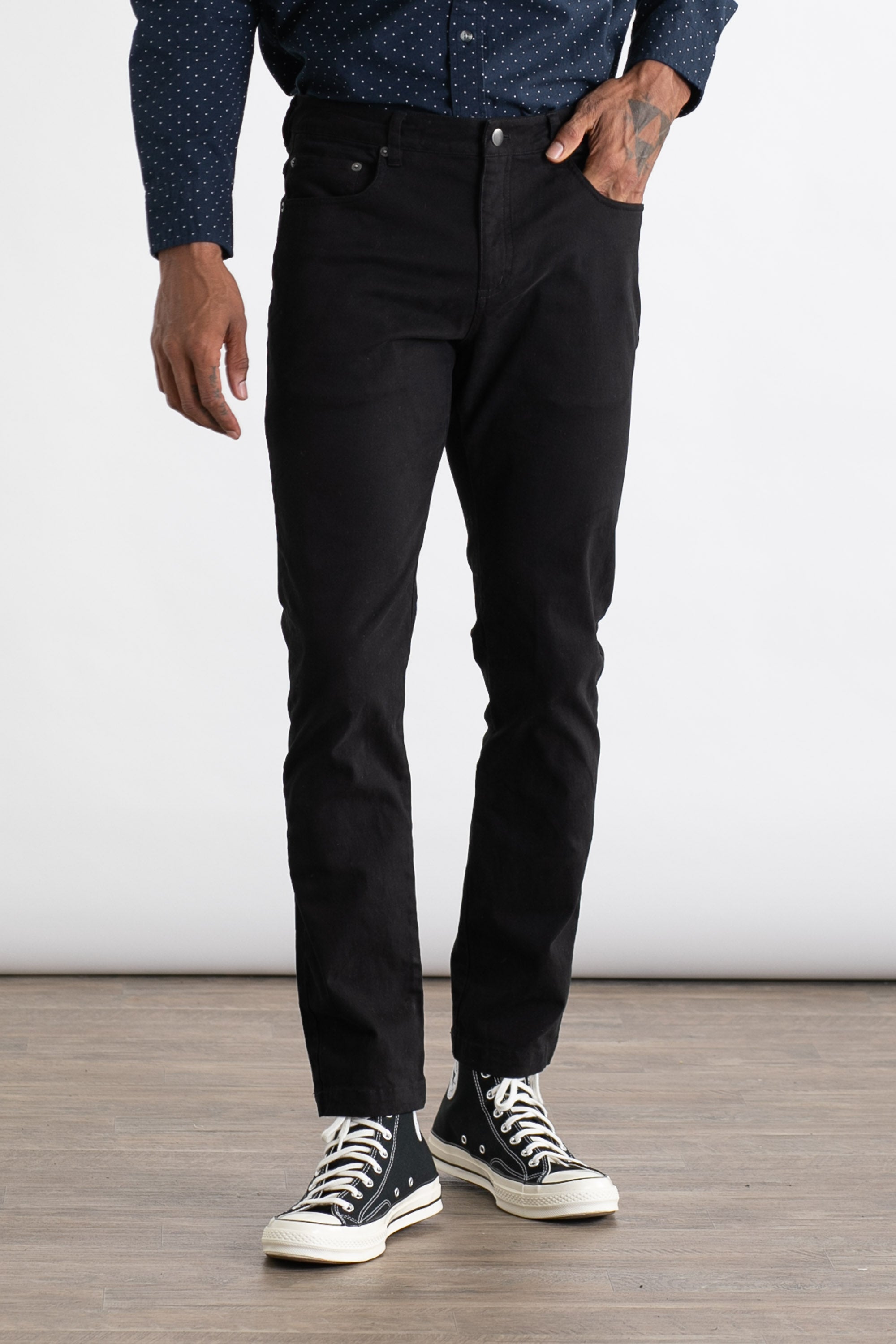 A person wears the Bridge & Burn Bradley Slim Pant in black, paired with a navy shirt with white dots and black high-top sneakers, standing on a wooden floor against a plain white background.