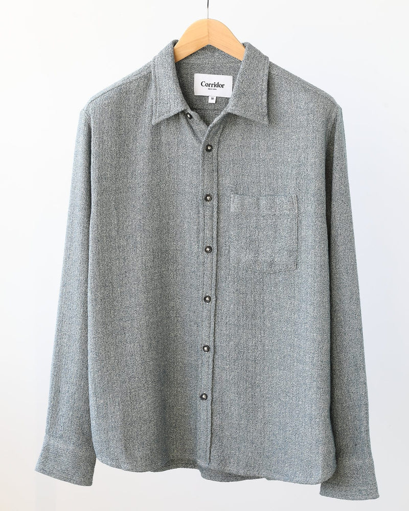 The Corridor Pearl Boucle Shirt in navy, a long-sleeved cotton shirt by Corridor, hangs on a wooden hanger against a plain white background.