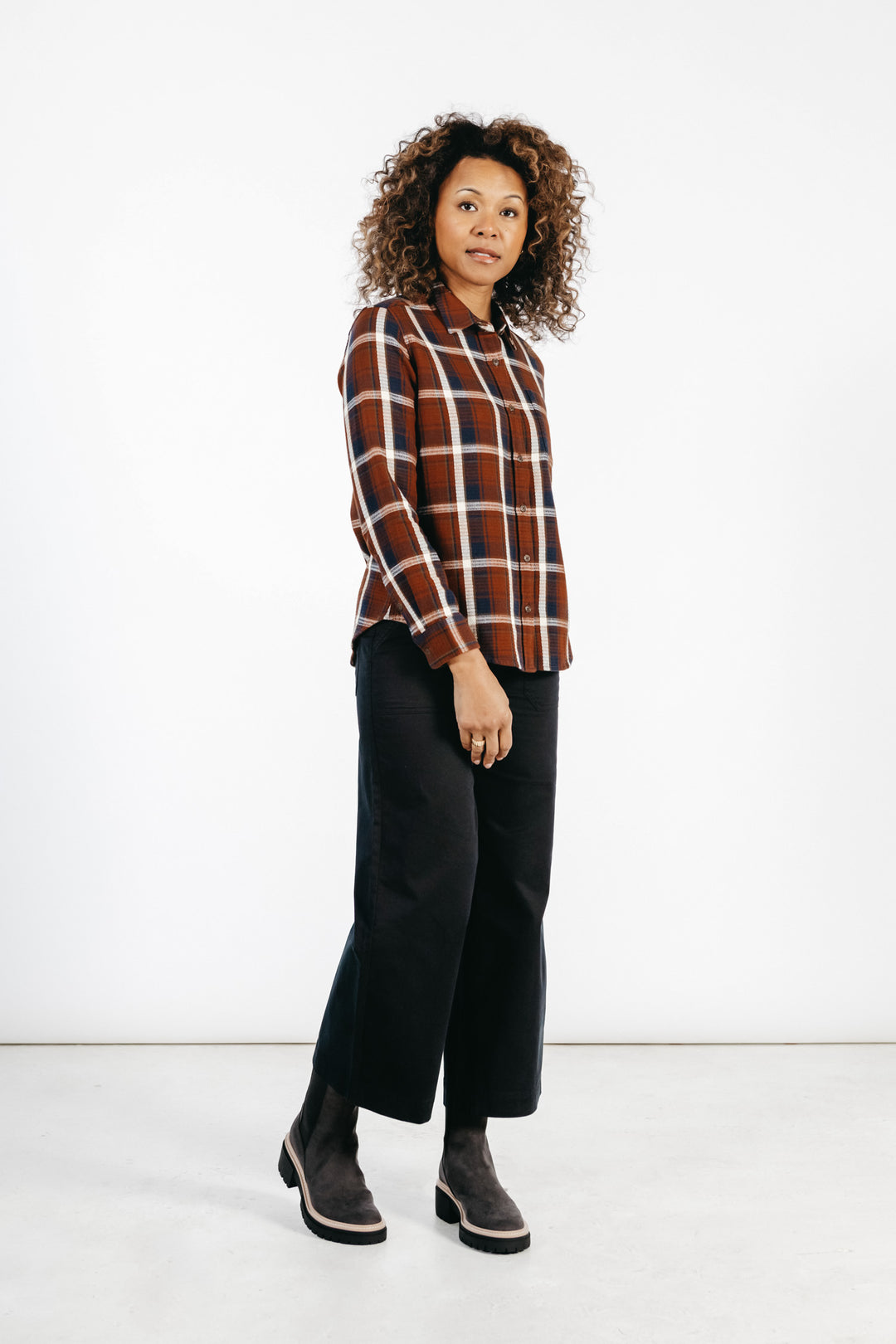 A person with curly hair wears the Bridge & Burn Ash Slim Shirt in Echo Ridge Plaid, paired with black wide-leg pants and dark boots, standing against a plain white background.