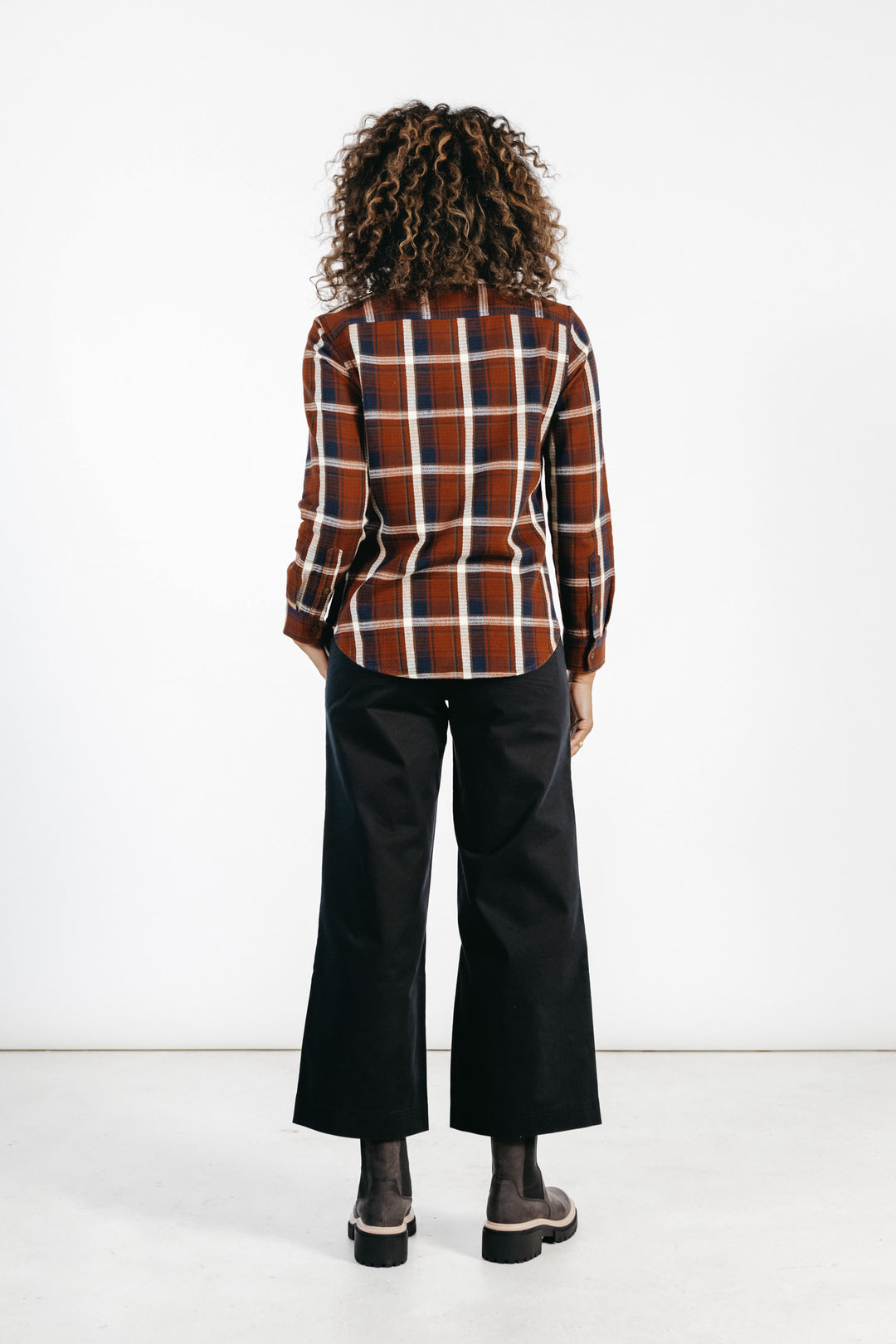 A person with curly hair faces away, wearing the Bridge & Burn Ash Slim Shirt in Echo Ridge Plaid—red and brown—paired with black wide-leg pants and dark shoes, set against a plain white background.