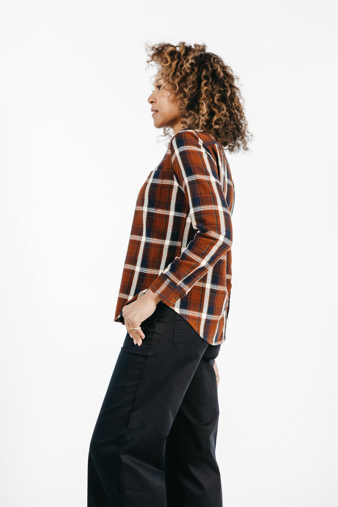 A woman with curly hair wears the Bridge & Burn Ash Slim Shirt in Echo Ridge Plaid and black pants, standing in profile against a plain white background.