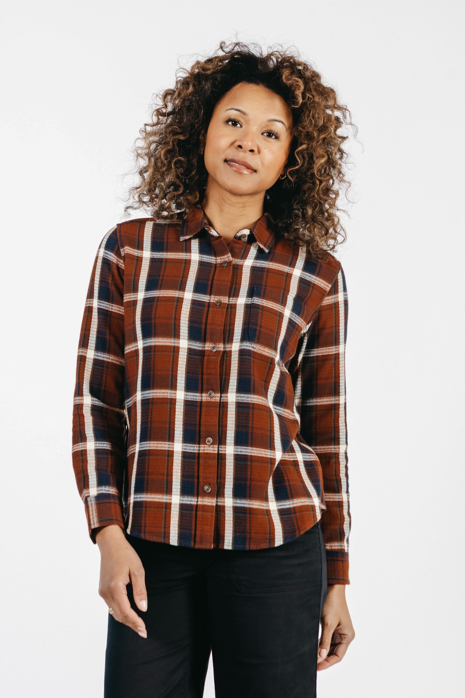 A woman with curly hair wears the Bridge & Burn Ash Slim Shirt in Echo Ridge Plaid, a slim-fit button-up in brown, white, and navy organic cotton. She pairs it with black pants and stands against a plain white background, looking relaxed at the camera.