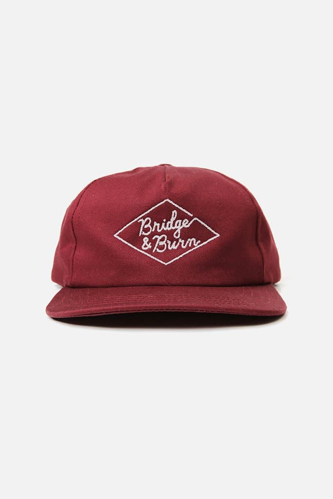 The Bridge & Burn Diamond Head Cap in Burgundy is a maroon baseball cap made from durable cotton twill, featuring white embroidered “Bridge & Burn” text inside a diamond-shaped outline on the front. It has a flat brim and a leather adjustable strap at the back for a comfortable fit.