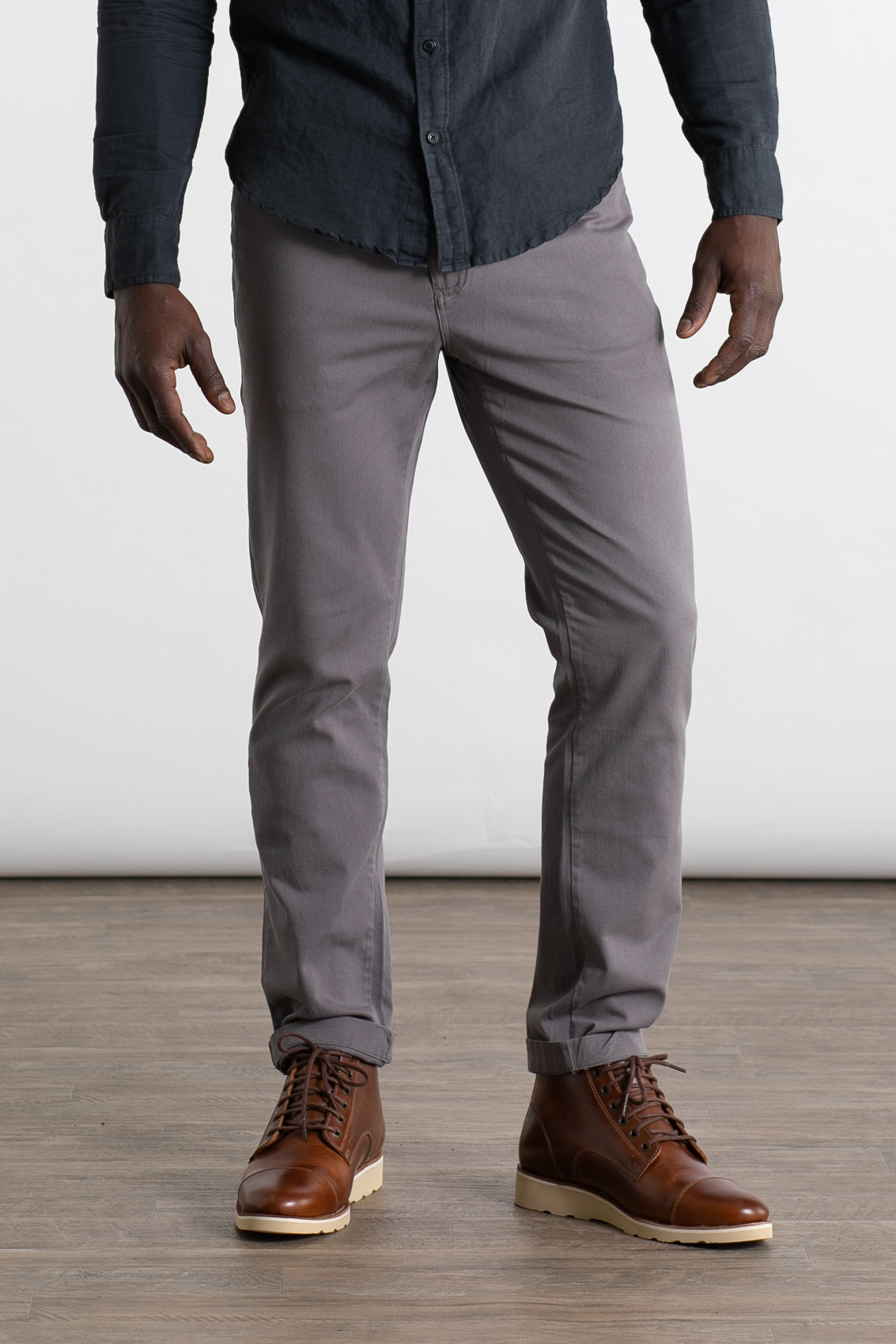 A person wearing a dark button-up shirt, Bridge & Burn Bradley Slim Pant in grey, and brown leather boots stands on a wooden floor against a plain white background.