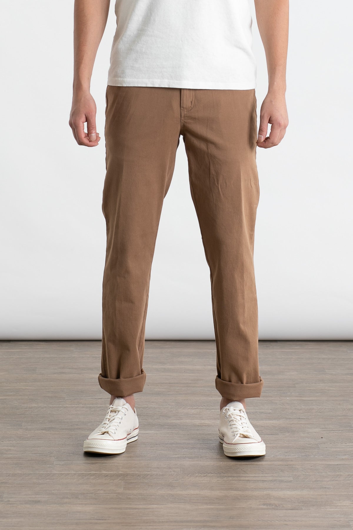 A person wearing a white t-shirt, white sneakers, and Bridge & Burn's Polk Pant in brown stands on a wooden floor against a plain white background.