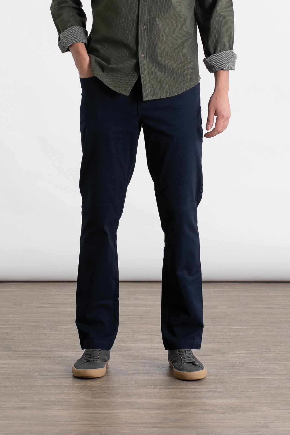 A person stands indoors on a wooden floor wearing Bridge & Burn’s Polk Pant in Navy, a green button-up shirt with rolled sleeves, and gray sneakers. The person's head is not visible in the image.