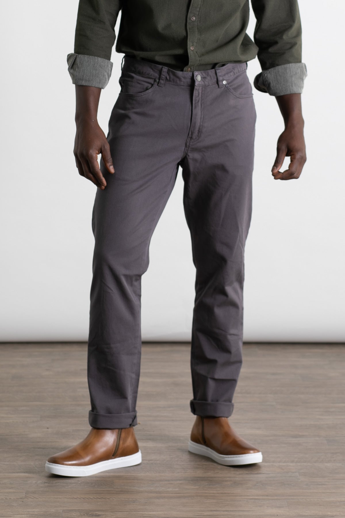 A person wearing a dark green shirt, Bridge & Burn's Polk Pant in Charcoal, and brown slip-on shoes stands on a wooden floor against a plain white background. The image is cropped at the shoulders.