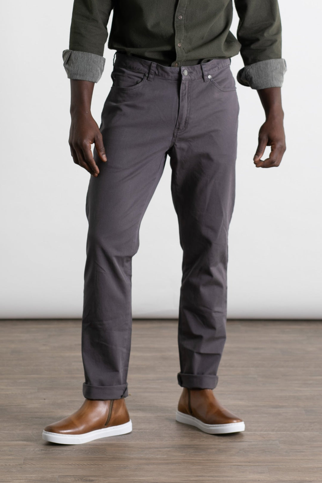 A person wearing a dark green shirt, Bridge & Burn's Polk Pant in Charcoal, and brown slip-on shoes stands on a wooden floor against a plain white background. The image is cropped at the shoulders.