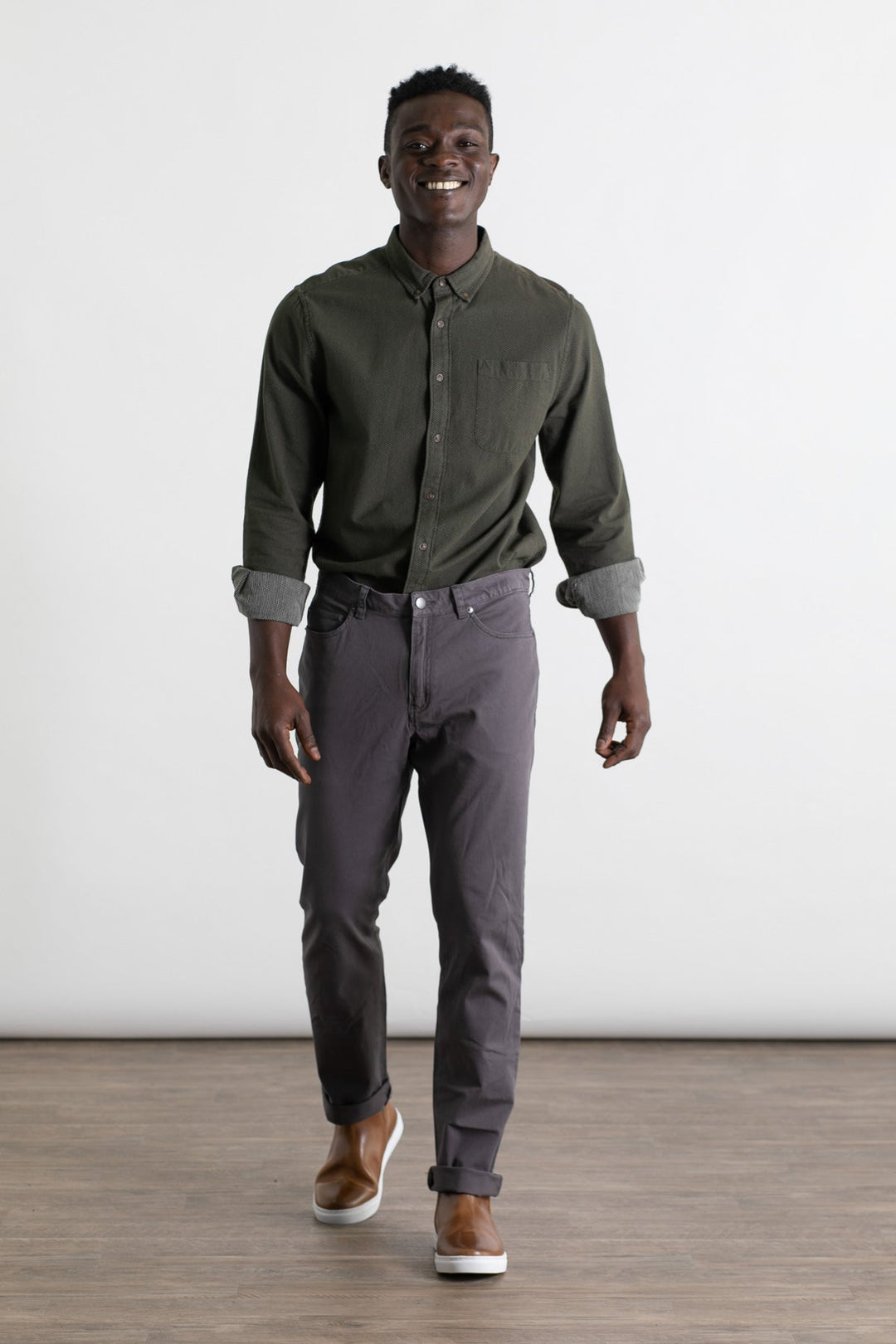 A man with short hair smiles while walking in a well-lit room, wearing the Bridge & Burn Polk Pant in Charcoal, a dark green button-up shirt, and brown slip-on shoes. The space features a wooden floor and plain white background.