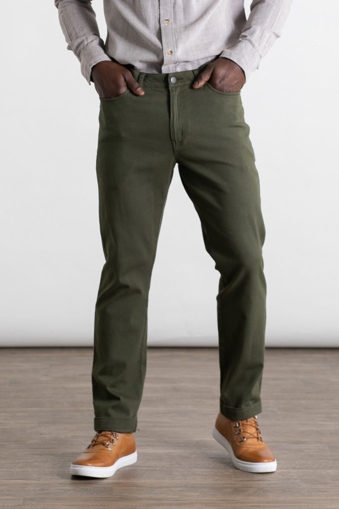 Wearing Bridge & Burn's Polk Pant in Olive, styled with a light gray button-up and tan sneakers, a person stands on a wooden floor with hands in pockets; the photo is cropped from shoulders to feet.