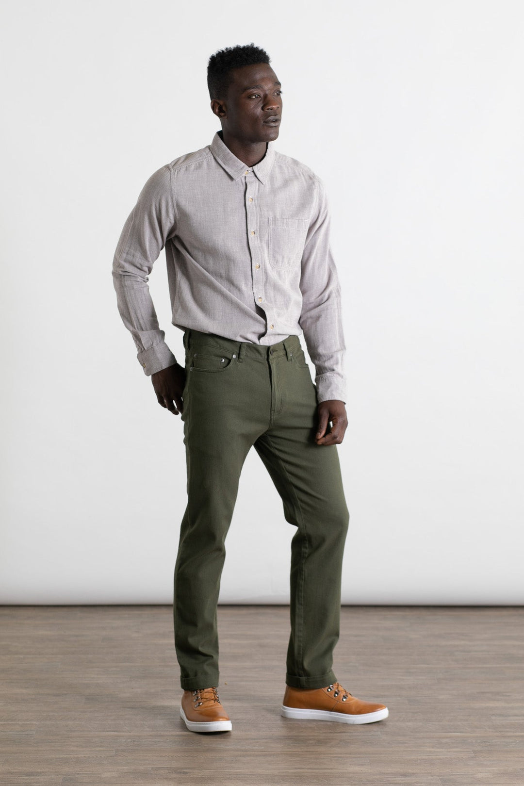 A man stands on a wooden floor against a white background, wearing Bridge & Burn's Polk Pant in Olive, a light gray button-up shirt, and tan sneakers. He faces slightly left with his hands by his sides.