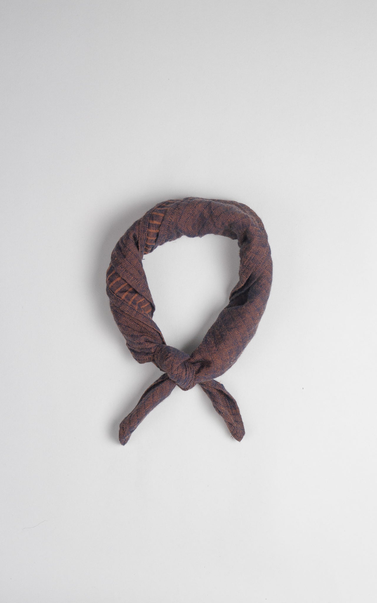 The Bridge & Burn Bandana / Subtle Stripe, made of organic cotton with a brown pattern, is tied in a loose knot and arranged in a circle on a plain light gray background.