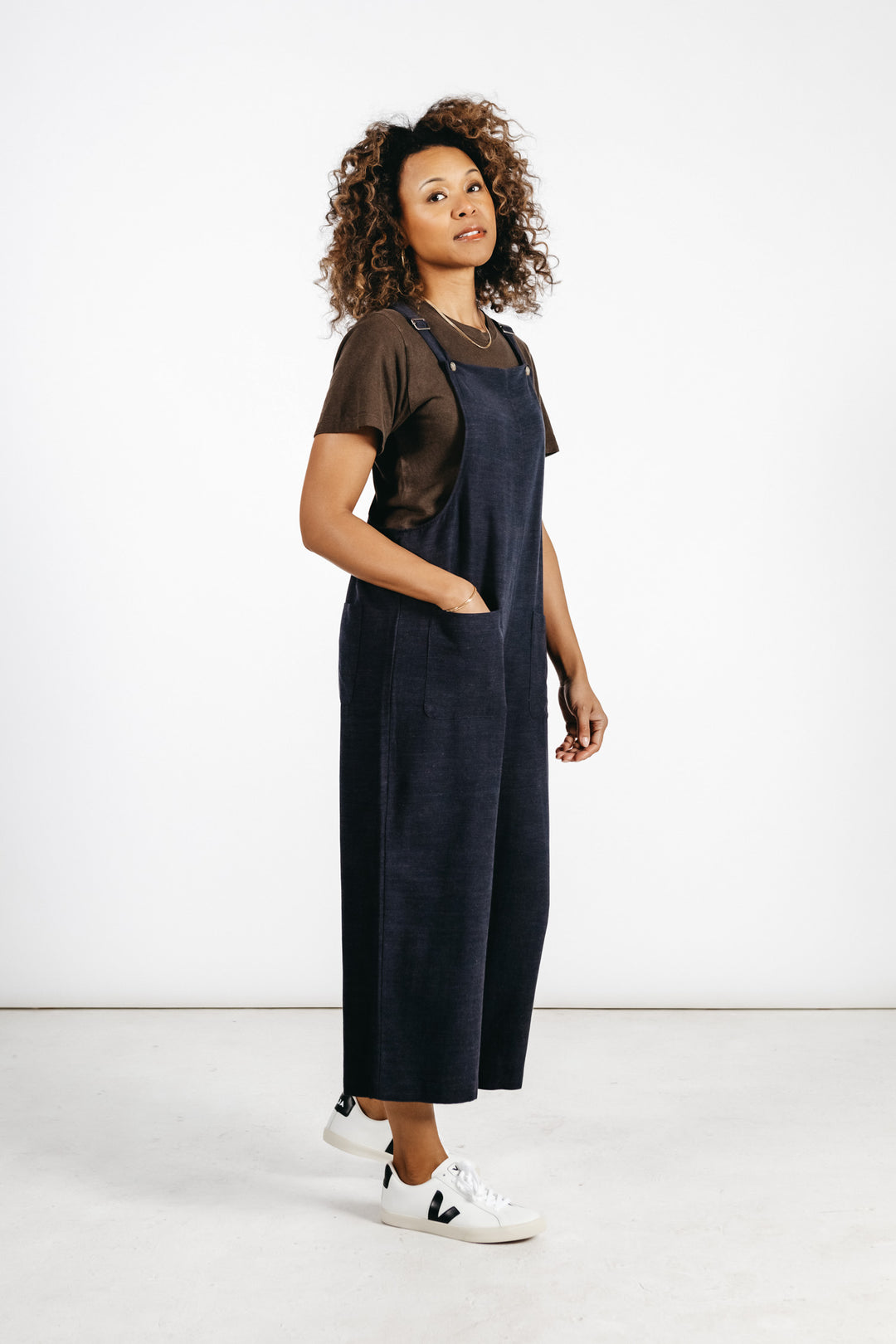 A woman with curly hair wears a brown t-shirt, white sneakers, and the Bridge & Burn Edie Overall in Navy—a wide leg linen-cotton blend style with adjustable straps. She stands in a minimal studio with a white wall and floor, hand in pocket, looking at the camera.