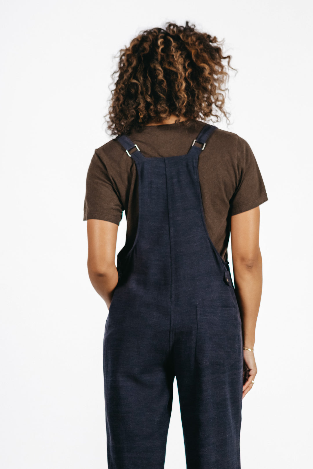 A person with curly hair, wearing a brown t-shirt and the Bridge & Burn Edie Overall in Navy—featuring wide legs and adjustable straps—is standing with their back to the camera against a plain white background.