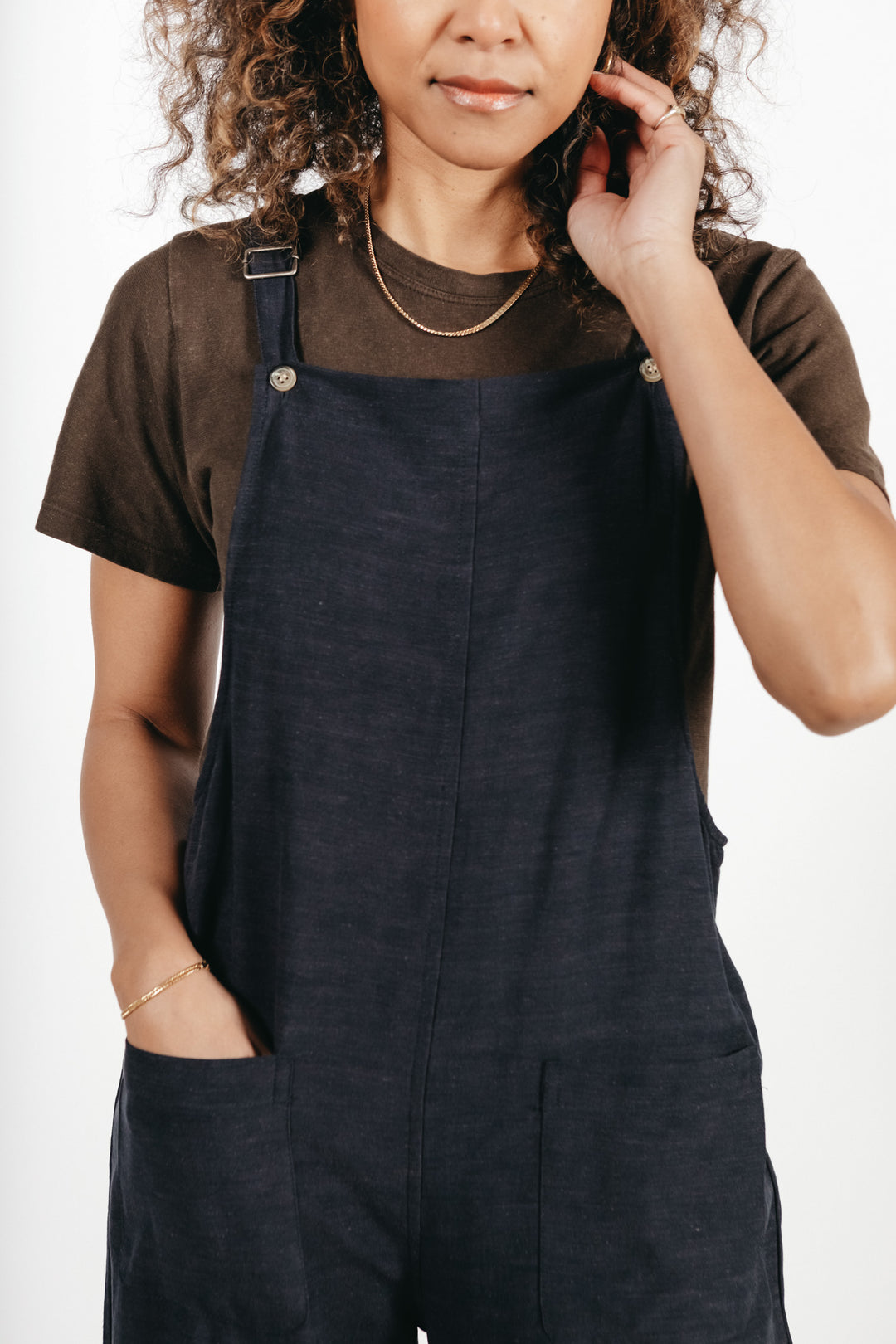 A woman stands facing the camera against a plain white background, wearing a dark t-shirt paired with the Bridge & Burn Edie Overall in Navy, featuring wide legs and adjustable straps; she has one hand in her pocket and the other touching her hair.