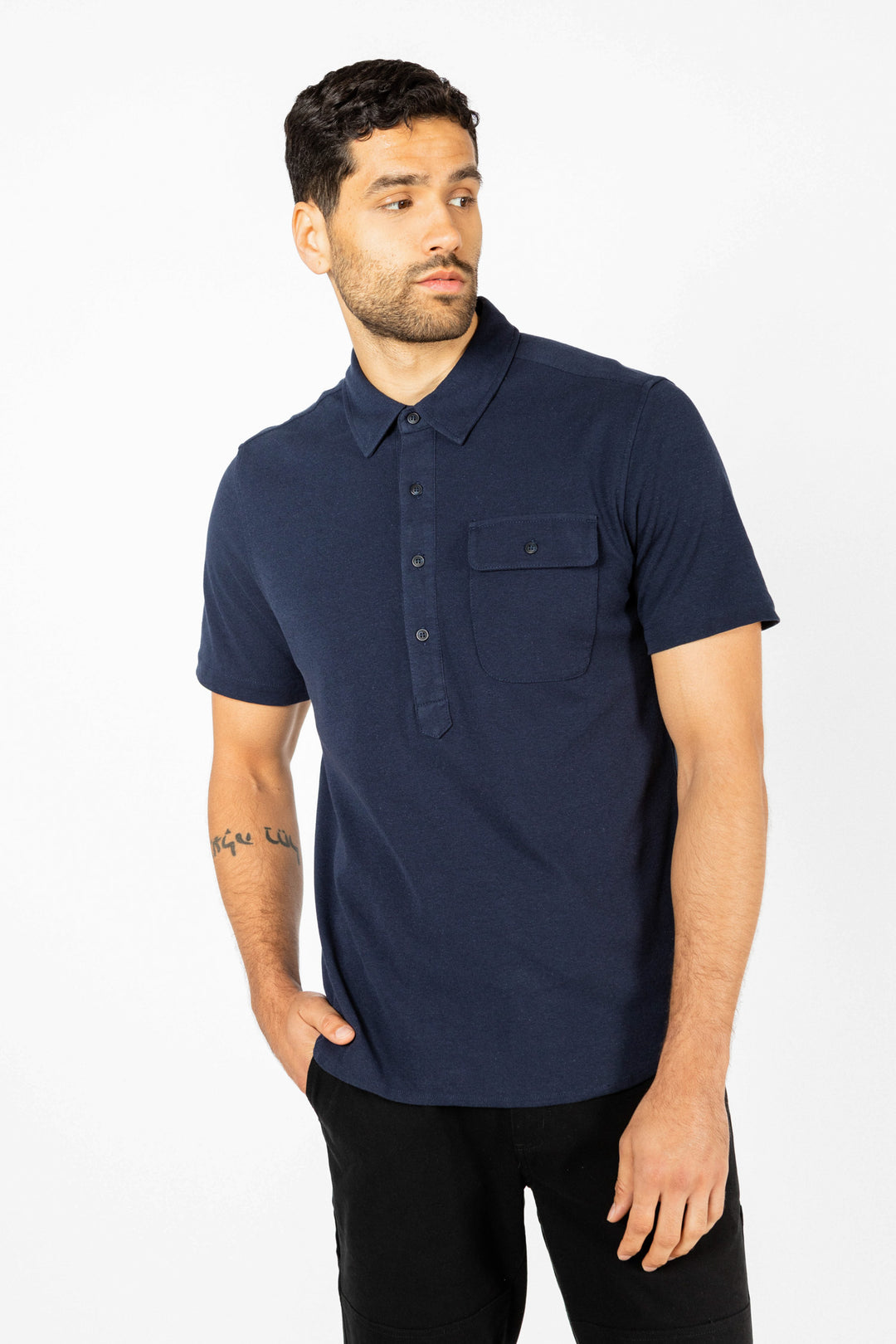 A man with short dark hair and a trimmed beard wears the Bridge & Burn Gary Knit Pullover in navy—a jersey knit, short-sleeved shirt with a chest pocket—paired with black pants, posing against a plain white background.