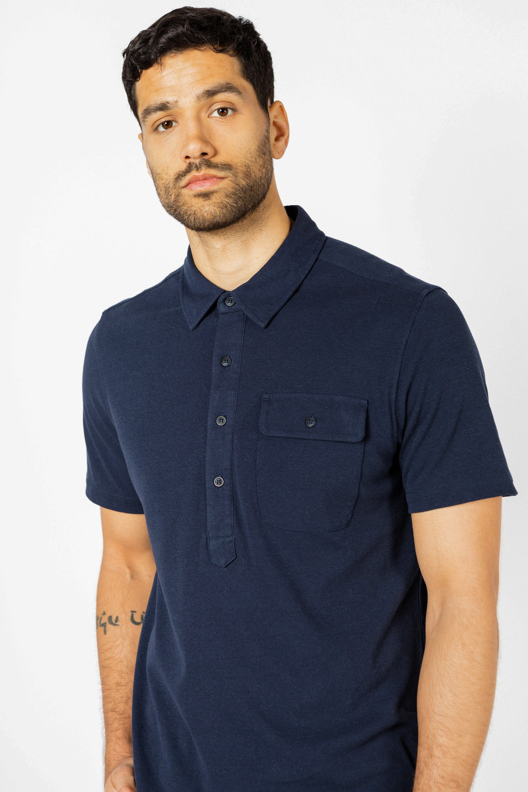 A man with short dark hair and a trimmed beard wears the Bridge & Burn Gary Knit Pullover in navy jersey, a men's summer short-sleeved shirt with a buttoned chest pocket, standing against a plain light background.