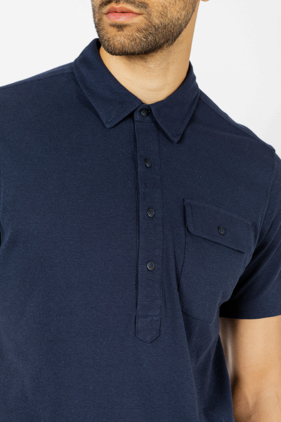 A man wears the Bridge & Burn Gary Knit Pullover in navy jersey, a short-sleeved top with a buttoned chest pocket, shown from shoulders to mid-chest against a white background.