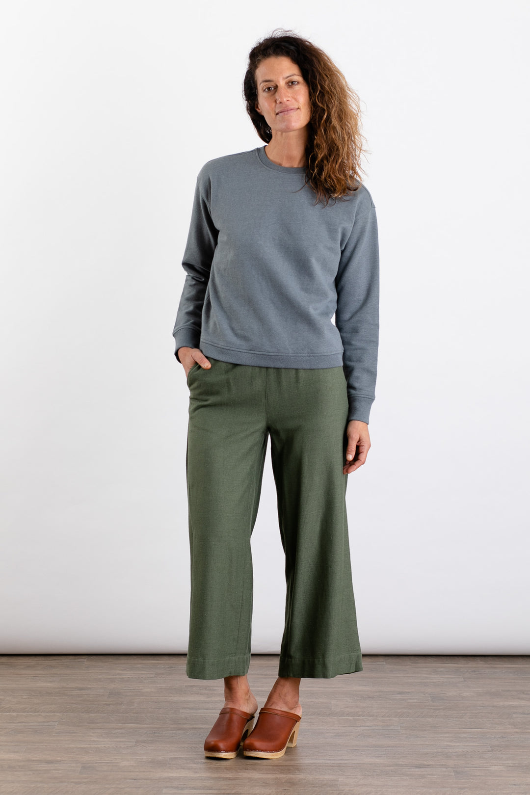 A woman with wavy brown hair stands against a white background, wearing the Bridge & Burn Hawthorne Boxy Crew Sweatshirt in Storm Blue, paired with olive green wide-leg pants and brown clogs, one hand in her pocket.