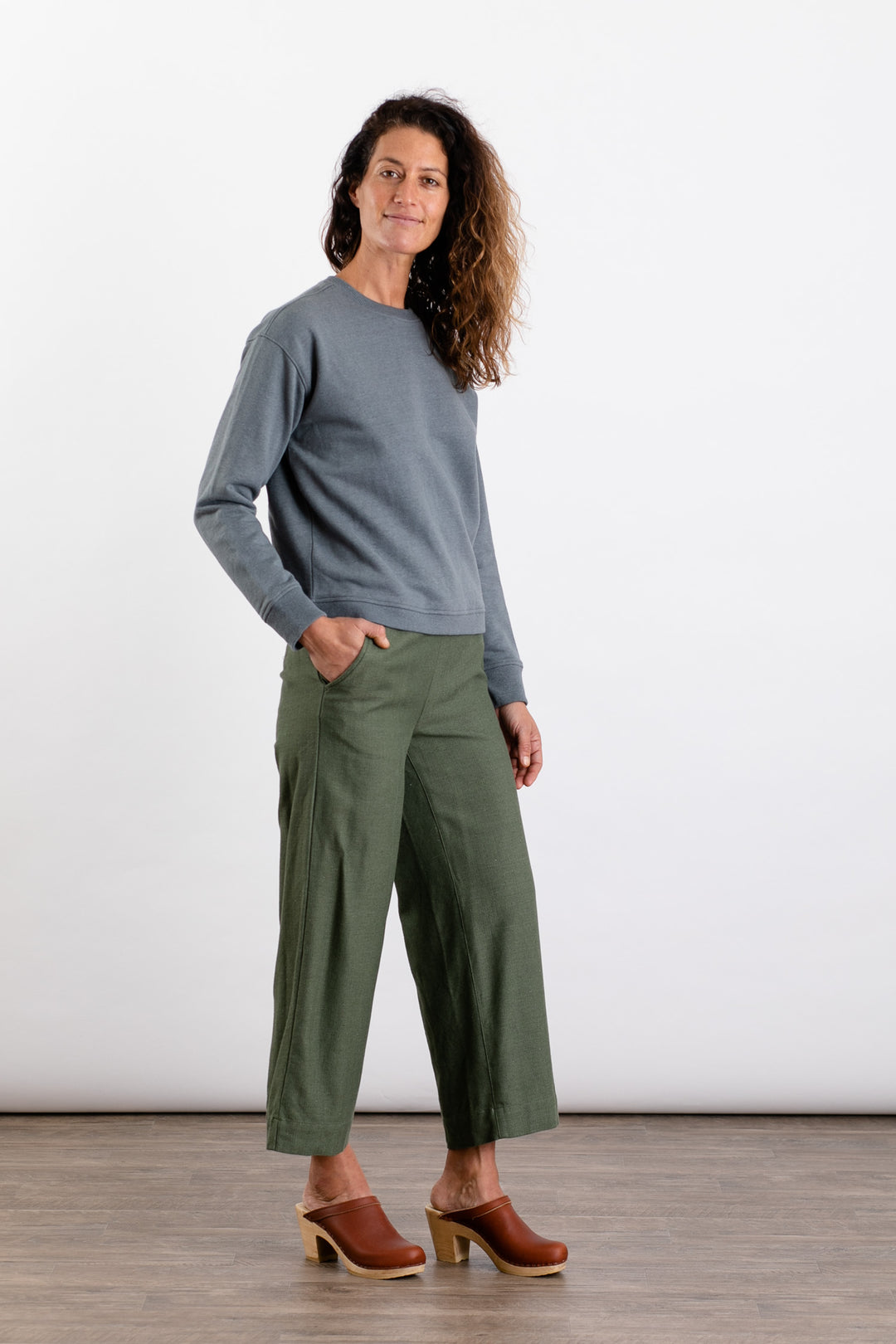 A woman with wavy brown hair smiles in a Bridge & Burn Hawthorne Boxy Crew Sweatshirt in Storm Blue, loose olive pants, and brown clogs. Her left hand is in her pocket as she poses against a plain white background.