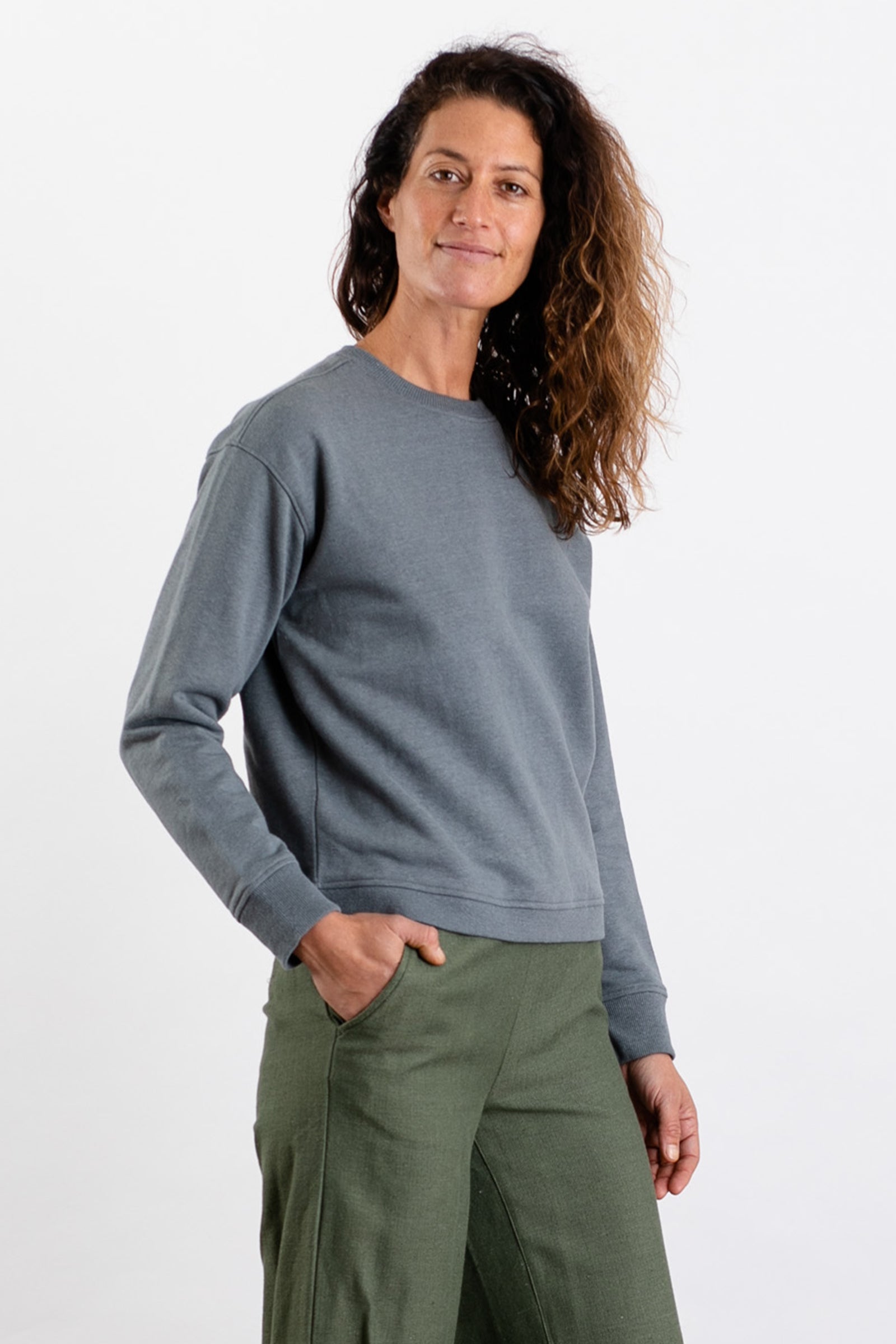 A woman with wavy brown hair wears the Bridge & Burn Hawthorne Boxy Crew Sweatshirt in Storm Blue, paired with olive green pants. She stands against a plain white background, smiling slightly at the camera with one hand in her pocket.