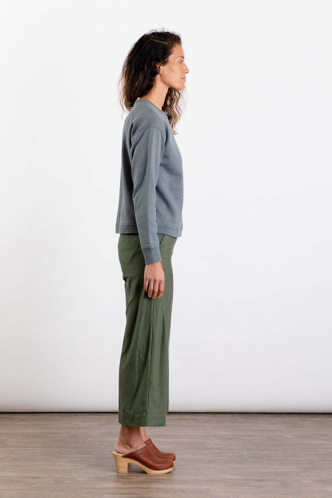 A woman stands in profile against a plain white background, wearing the Bridge & Burn Hawthorne Boxy Crew Sweatshirt in Storm Blue, paired with olive green wide-leg pants and brown wooden-heeled clogs.