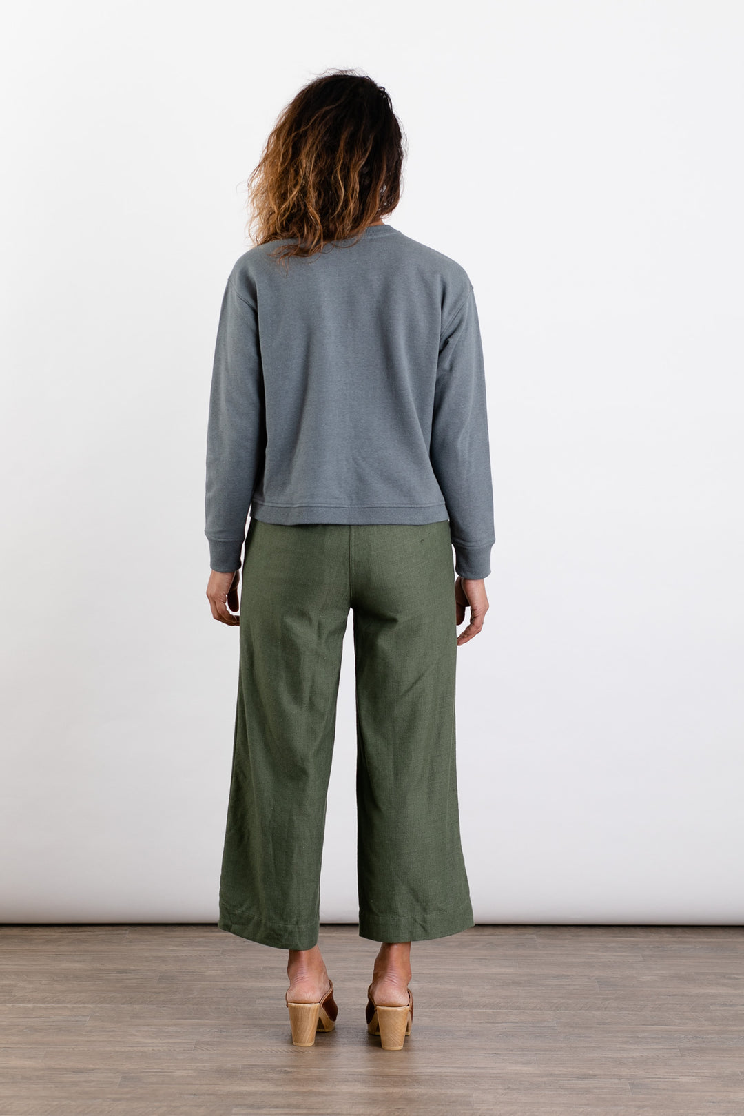 A person stands indoors with loose, wavy hair, wearing the Bridge & Burn Hawthorne Boxy Crew Sweatshirt in Storm Blue, olive green wide-leg pants, and tan sandals against a plain white background.