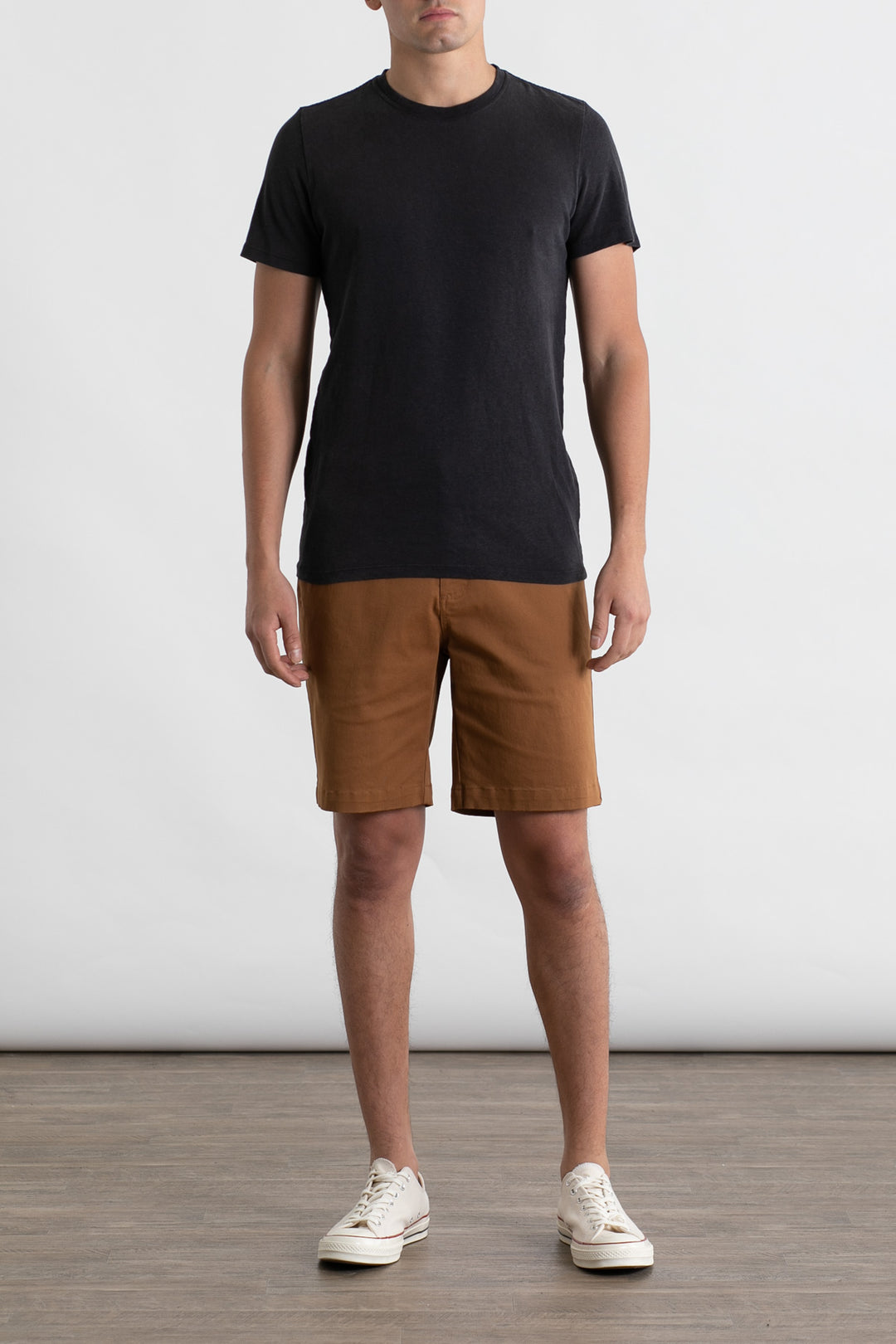 A person stands indoors on a wooden floor, wearing the Jungmaven Baja Tee in black by Jungmaven, brown knee-length shorts, and white sneakers. The background is a plain light gray wall and their face is not visible.