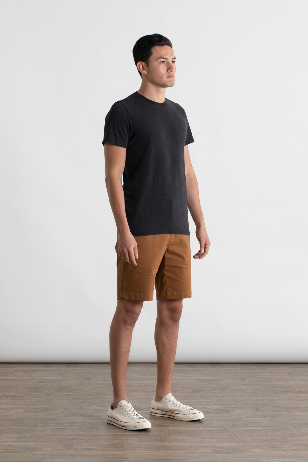 A man stands indoors on a wooden floor, wearing the Jungmaven Baja Tee in black by Jungmaven, brown shorts, and white sneakers, facing slightly to the right against a white background.