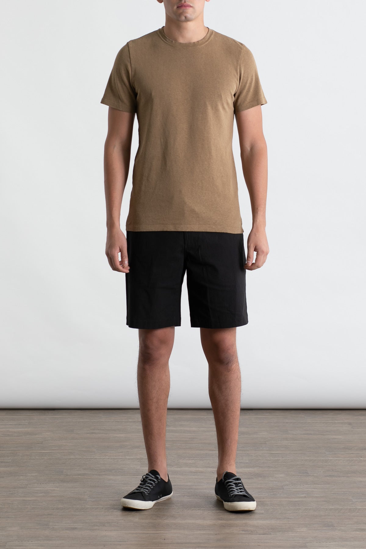 A person stands indoors against a plain light background, arms relaxed at their sides, wearing the Jungmaven Baja Tee in Coyote and black shorts with black sneakers featuring white soles.