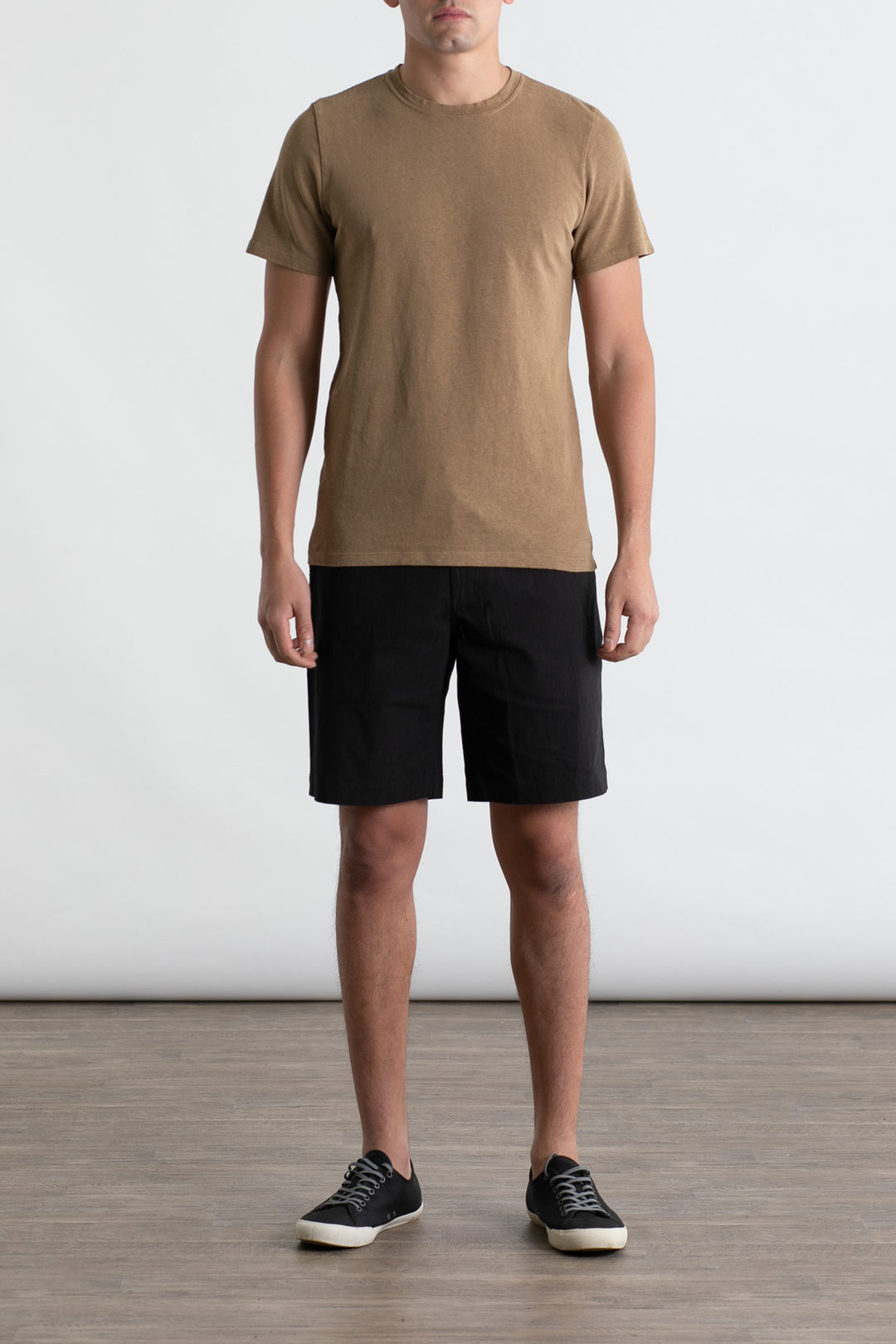 A person stands indoors against a plain light background, arms relaxed at their sides, wearing the Jungmaven Baja Tee in Coyote and black shorts with black sneakers featuring white soles.