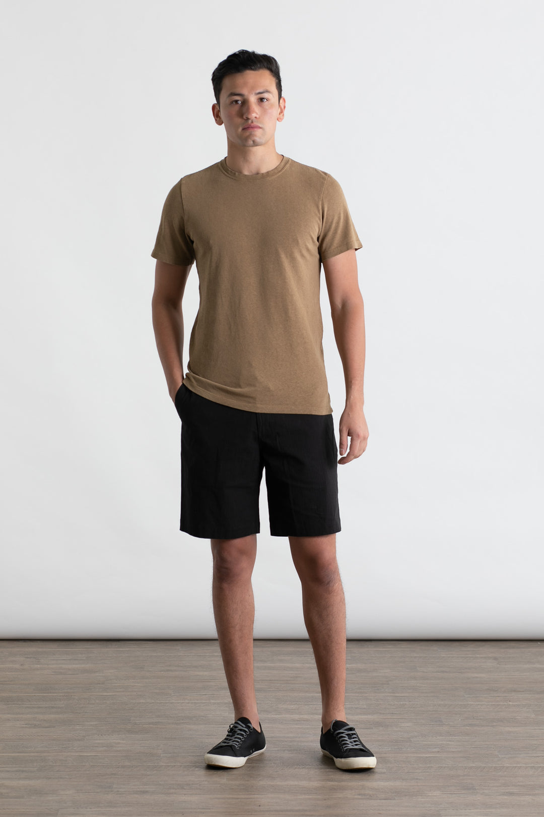 A man stands indoors against a plain white background, wearing the Jungmaven Baja Tee in Coyote. He pairs it with black shorts and black sneakers with white soles, has a neutral expression, and his hands rest at his sides.