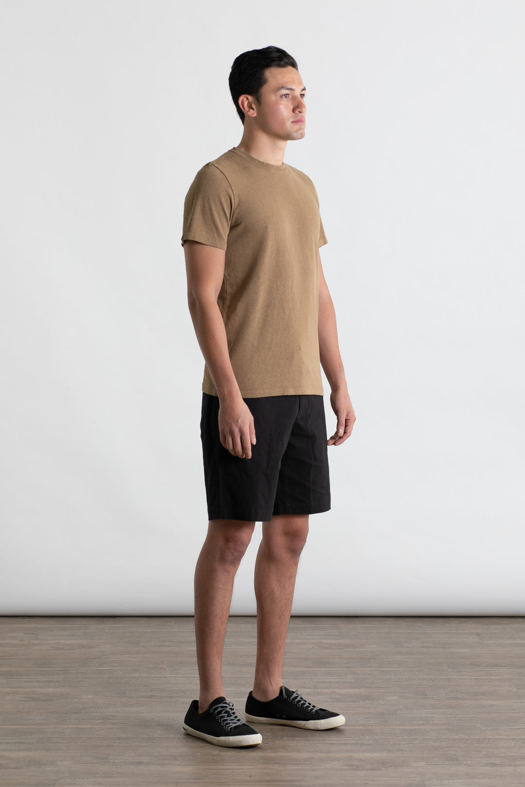A man stands indoors on a wooden floor against a white backdrop, wearing the Jungmaven Baja Tee in Coyote and black shorts with black sneakers. He is looking slightly to the side.