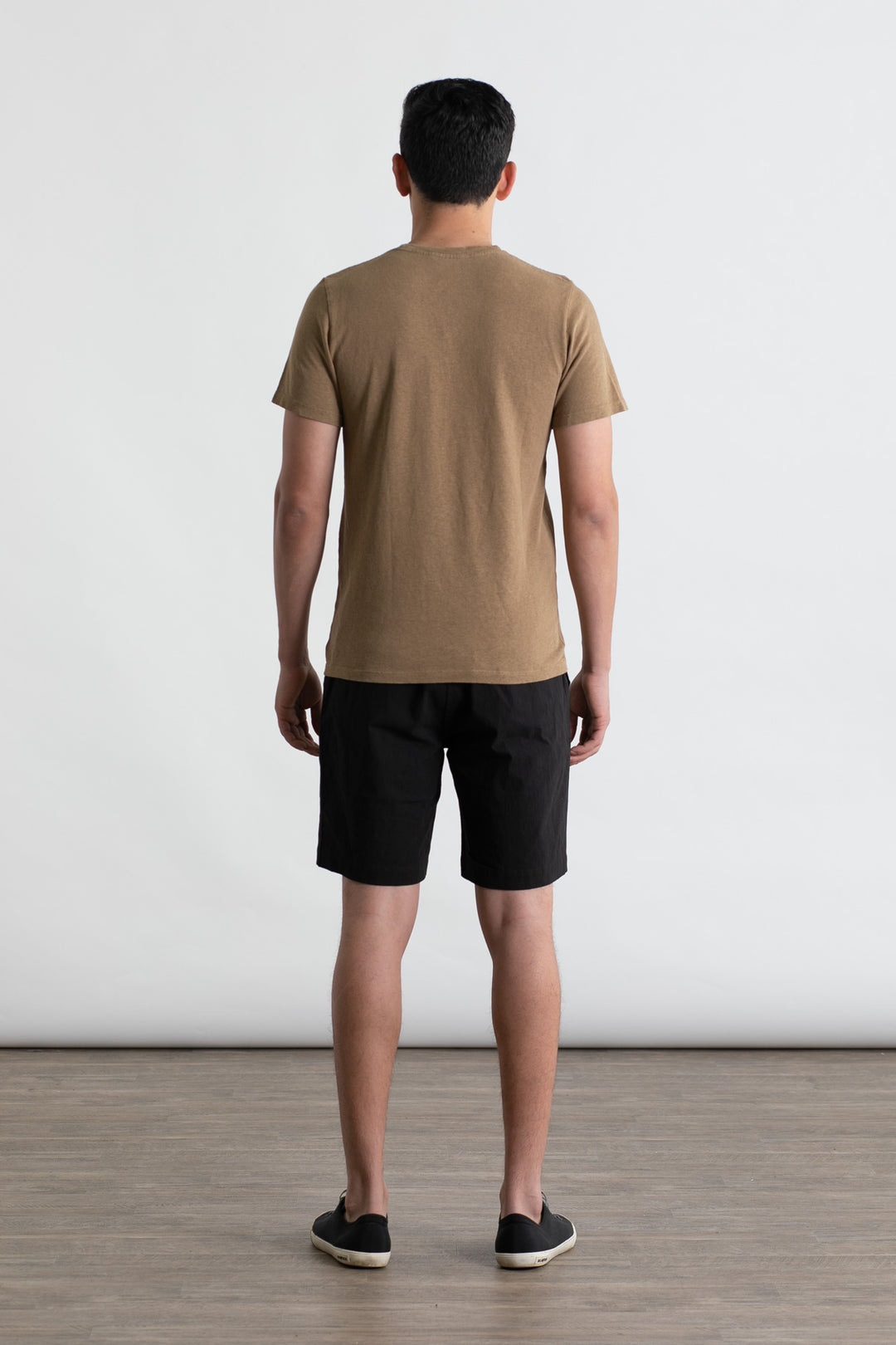 A man, facing away from the camera, wears a Jungmaven Baja Tee in Coyote with black shorts and black slip-on shoes. He stands on a wooden floor against a plain white background.