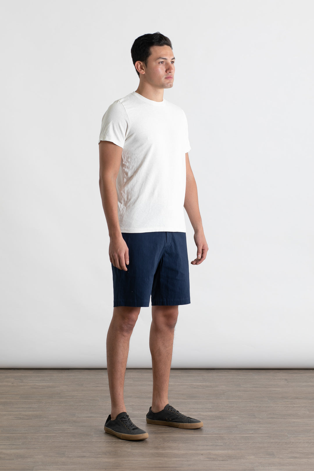 A man stands indoors on a wooden floor against a plain light background, wearing the Jungmaven Baja Tee in Washed White with navy blue shorts and dark sneakers. He faces slightly to the side with his arms relaxed by his sides.